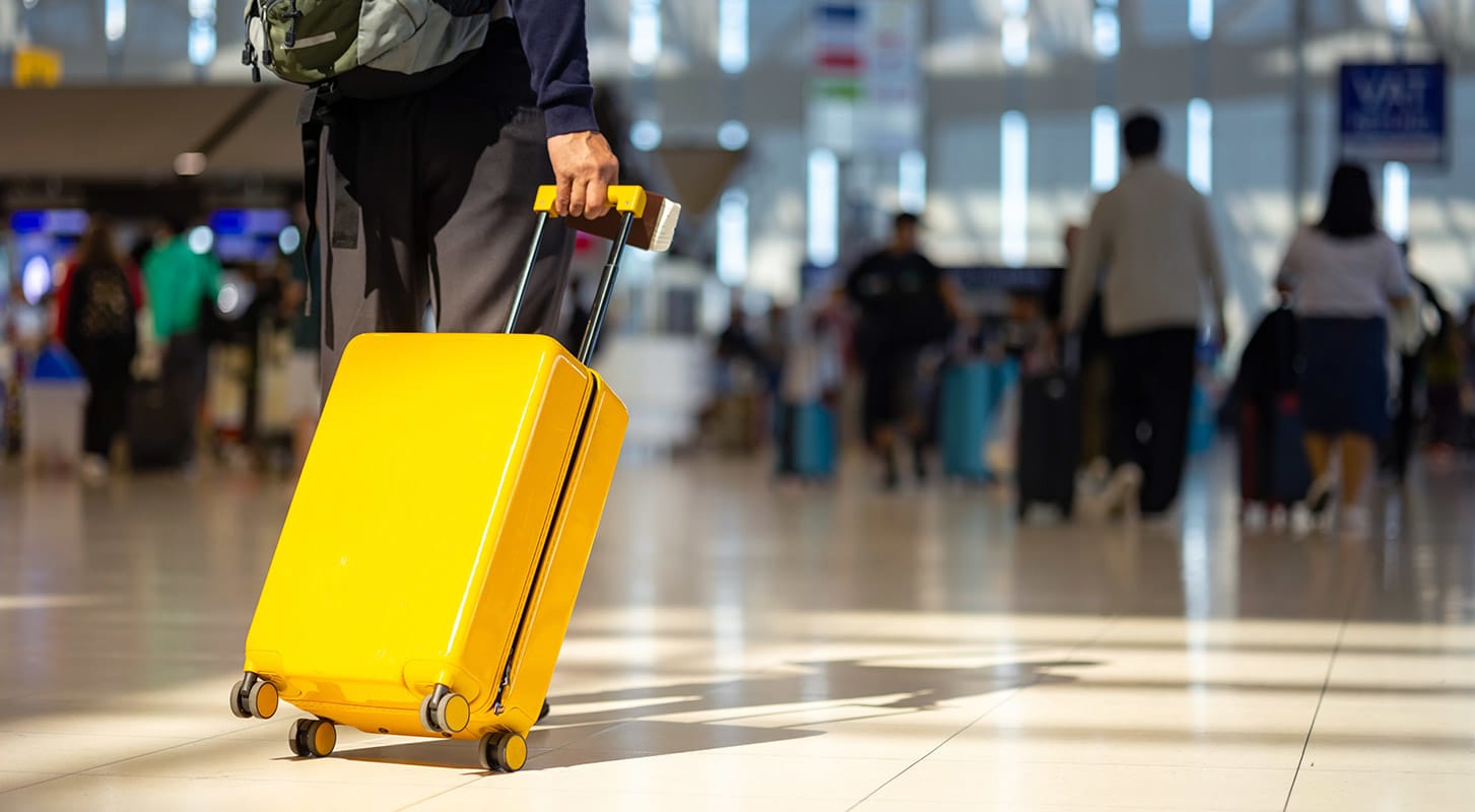 A traveler with a yellow suitcase stands in an airport terminal, surrounded by fellow passengers and departure signs.